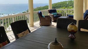 A covered outdoor seating area with wicker furniture and a table, overlooking a scenic view of the ocean and greenery.