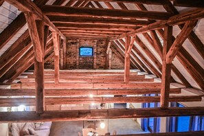 A rustic attic room with exposed wooden beams, a brick fireplace, and a television.