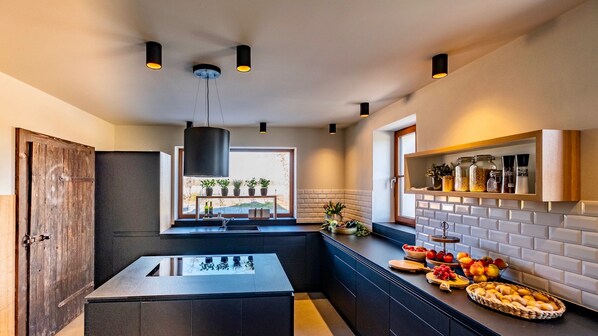 A modern kitchen with a central island, black pendant lights, a window with plants, and a brick backsplash.
