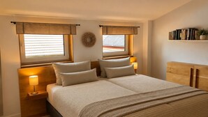 Une chambre à coucher avec un lit, des tables de chevet, une étagère fixée au mur avec des livres et une fenêtre avec des rideaux.