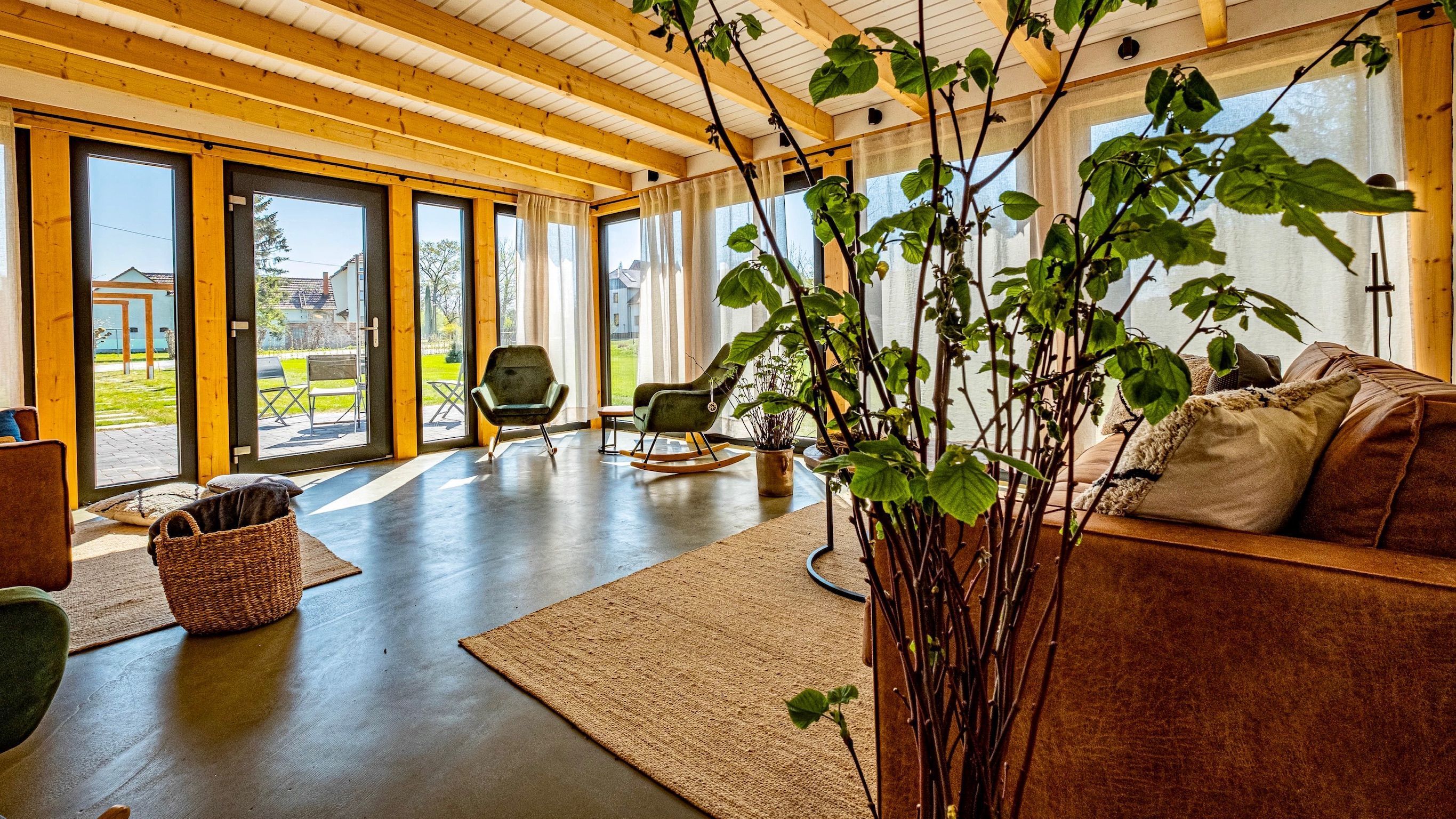 A spacious living room with a wooden ceiling, large windows, a rocking chair, a sofa, and a potted plant.