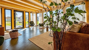 Amplia sala de estar con techo de madera, ventanas grandes, una mecedora, un sofá y una planta en maceta.