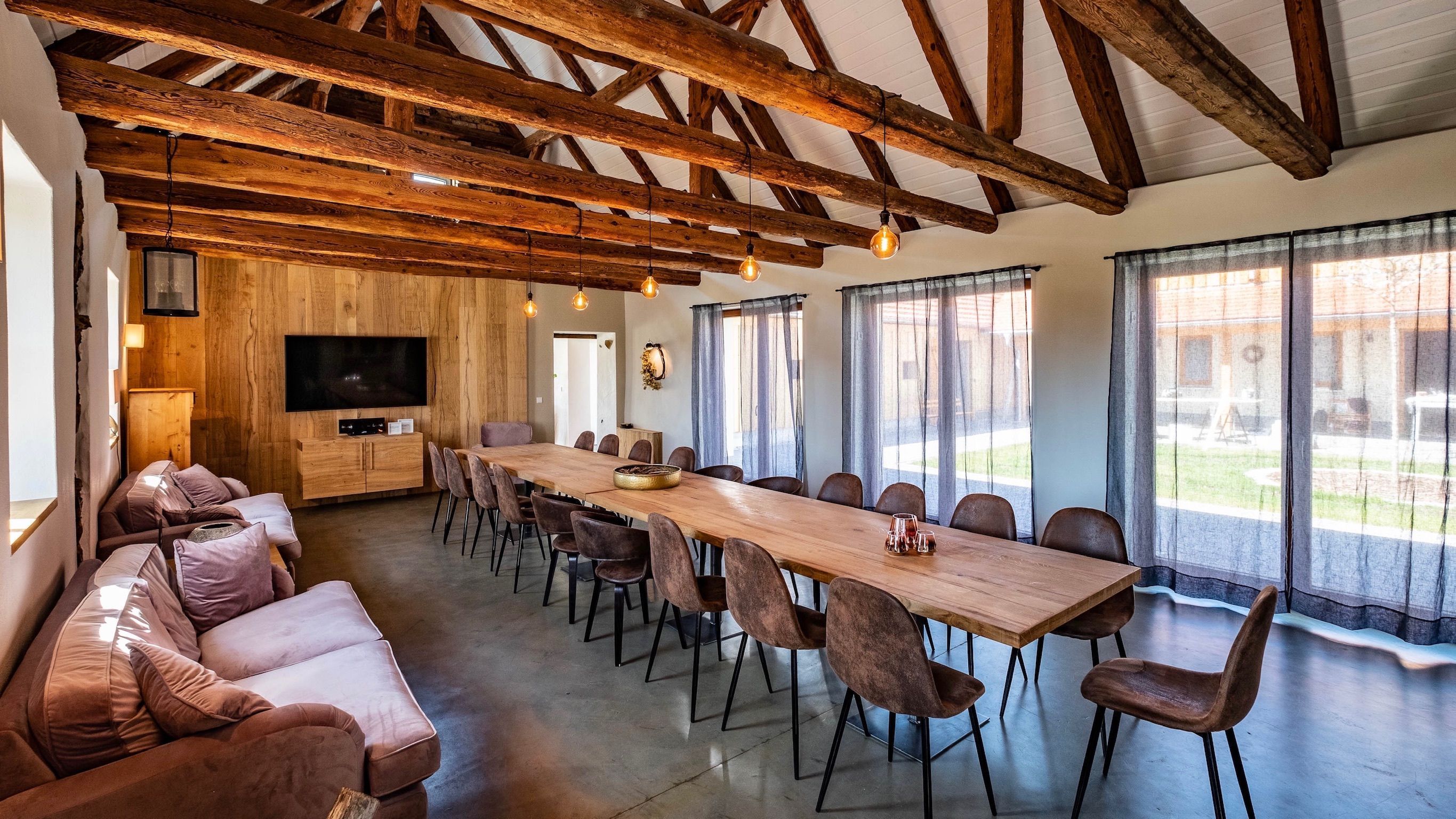 A spacious room with a long wooden table, cushioned seating, a flat-screen TV, and large windows.