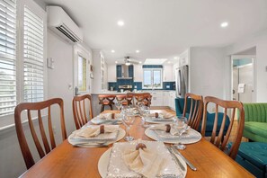 A dining area with a wooden table set for six, a kitchen in the background, and a green sofa.