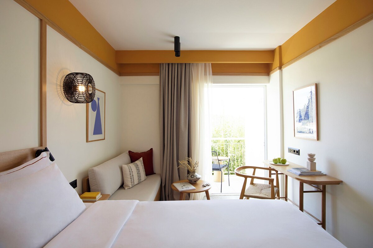 Ein Hotelzimmer mit einem großen Bett, einem kleinen Tisch mit Stuhl, einem Balkon mit Aussicht und einem Deckenventilator.