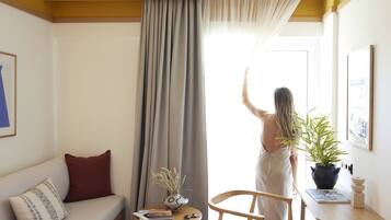 Una habitación con un sofá, una mesita con un florero y libros, una silla y un gran ventanal con cortinas.