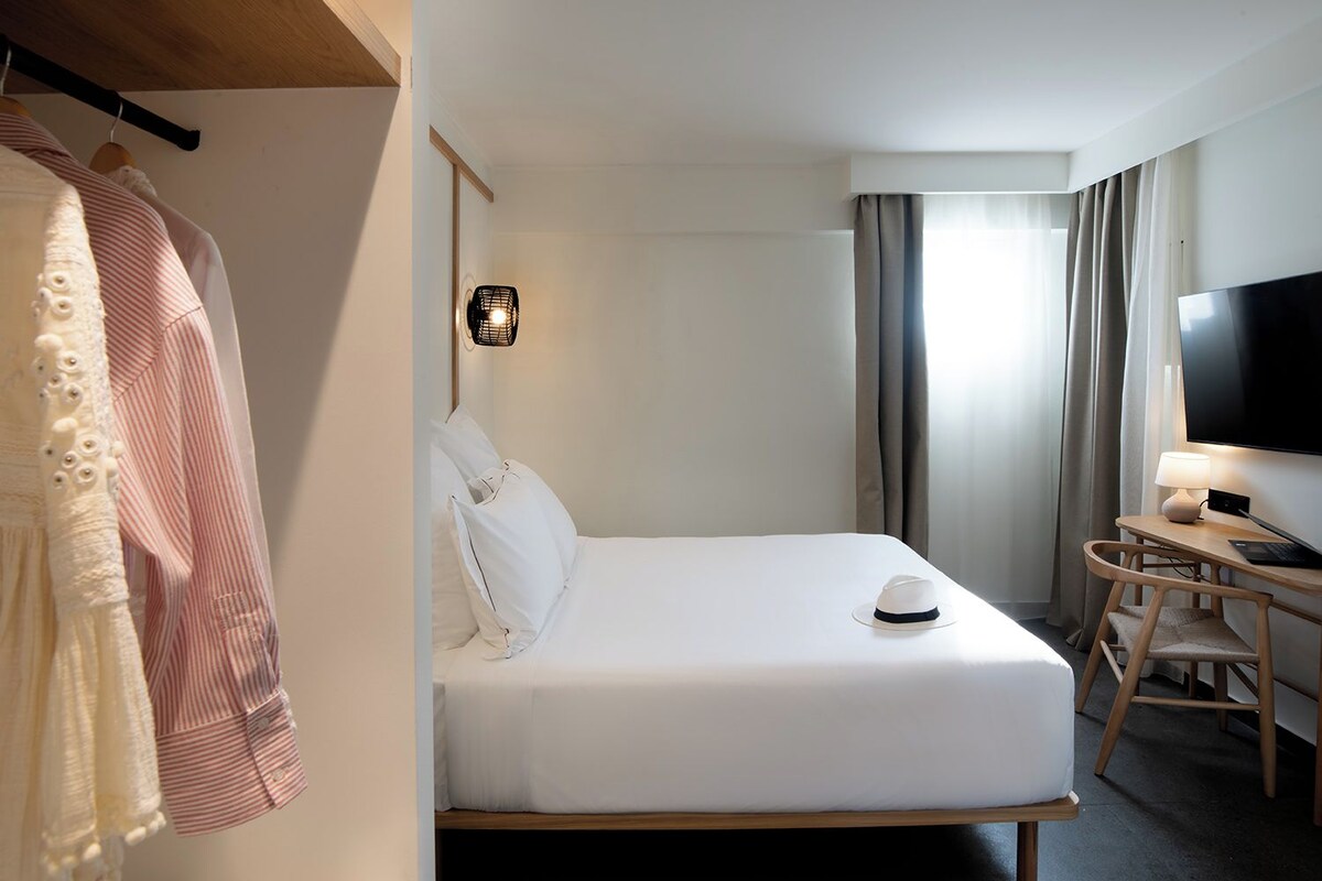 A hotel room with a bed, a desk with a chair, a TV, and a wardrobe.