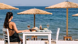 A woman dining at a table by the sea with thatched umbrellas and wooden chairs. - Brown Beach Evia Island, a member of Brown Hotels (Eretria)