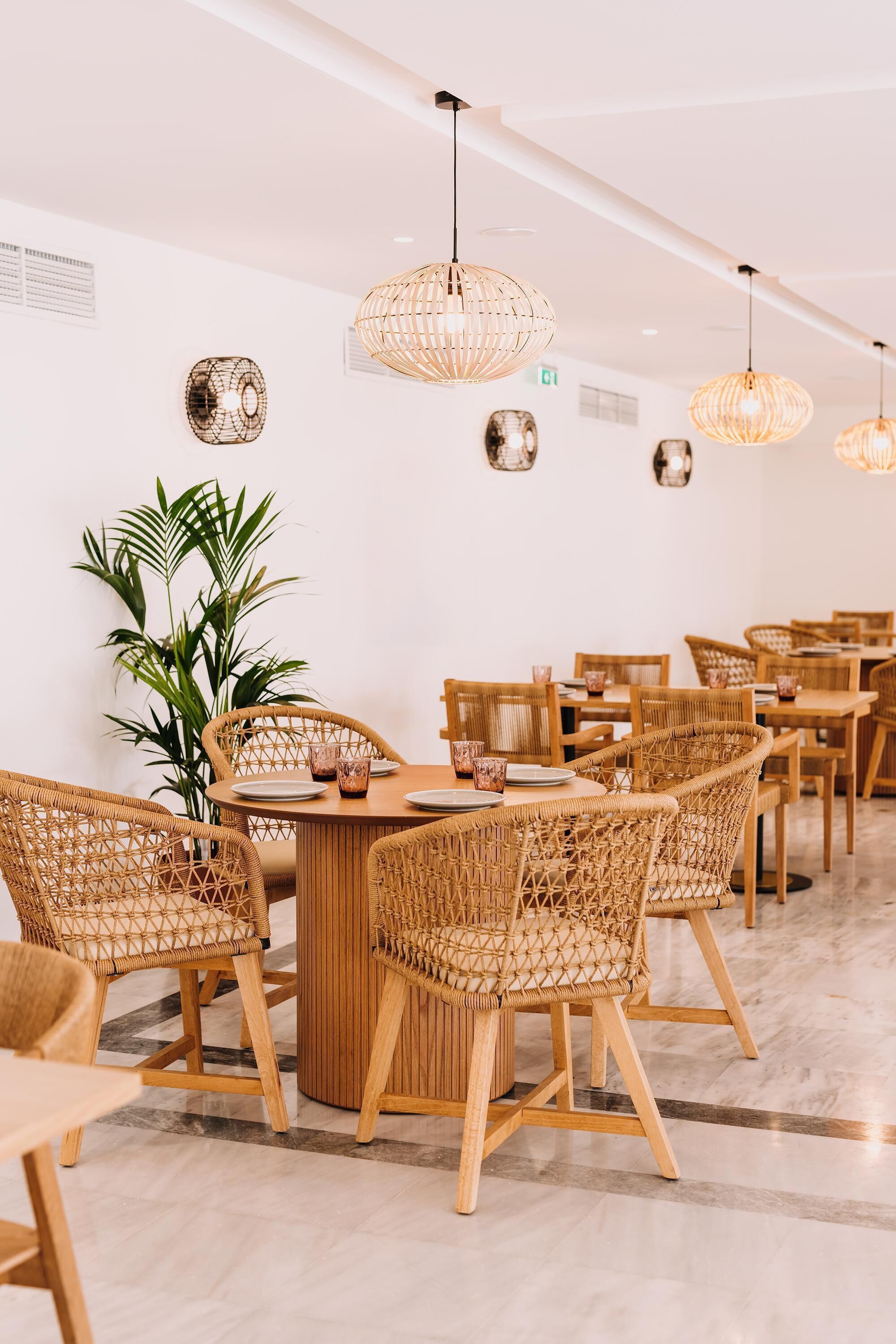 Un comedor con sillas de mimbre, mesas redondas y lámparas colgantes.
