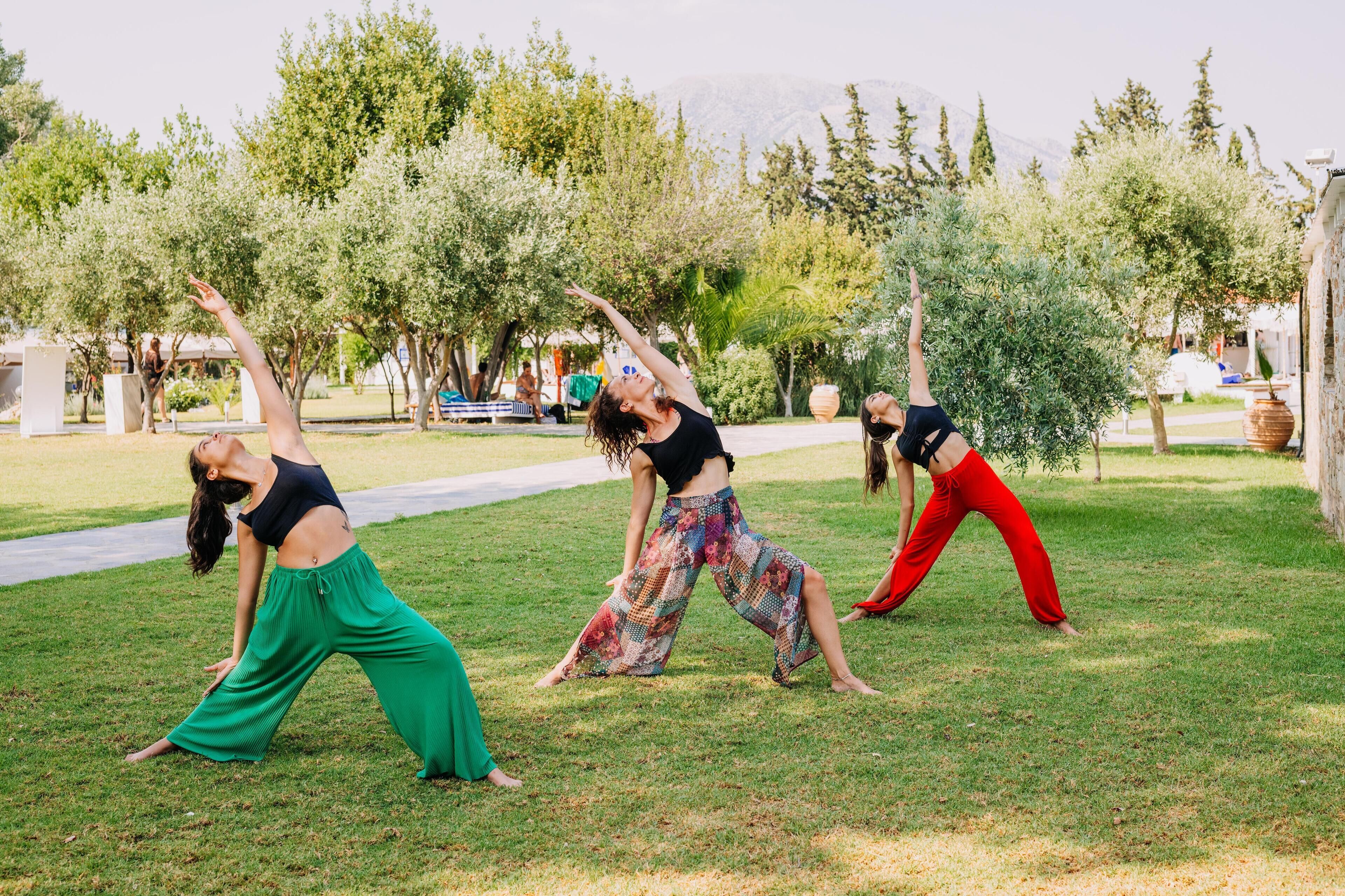 Trois personnes pratiquent le yoga en plein air, sur une pelouse, au milieu d’arbres, à proximité d’un sentier.