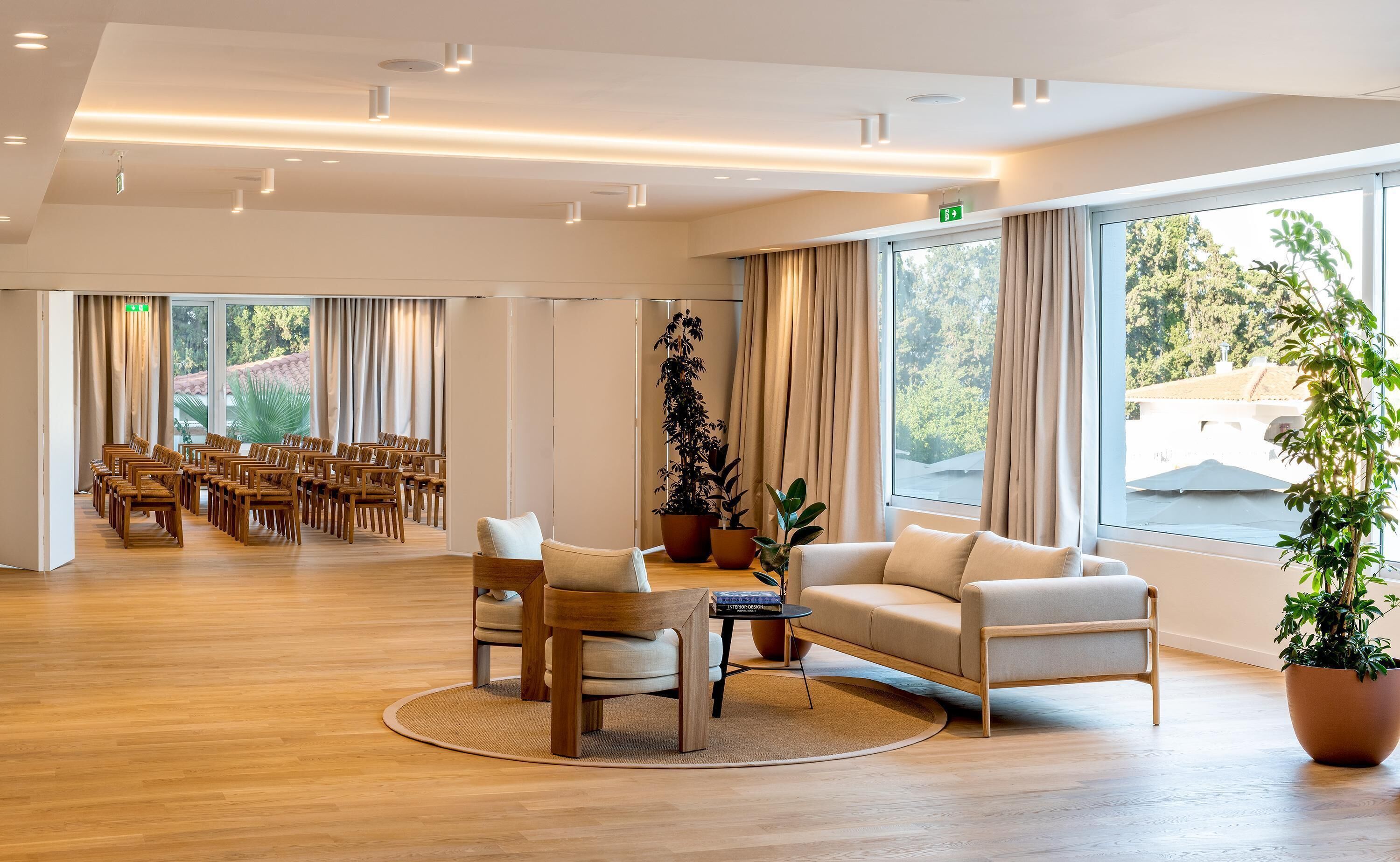 A spacious room with a wooden floor, a seating area with sofas and chairs, large windows with curtains, and potted plants.