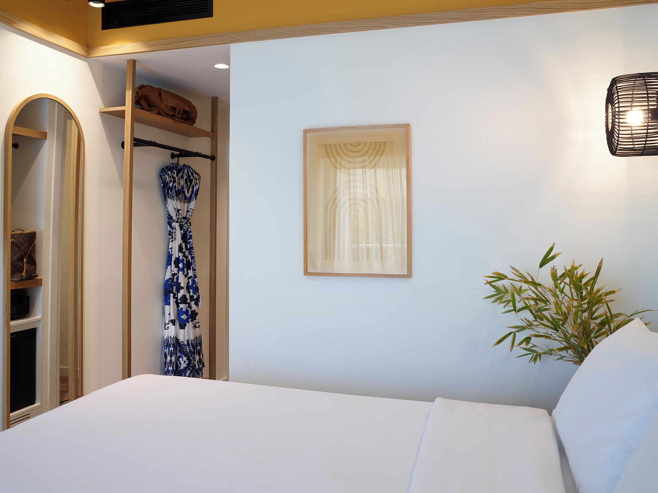 A bedroom with a bed, a wardrobe, a mirror, a framed picture, and a plant.