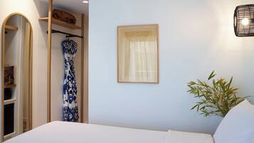 A bedroom with a bed, a wardrobe, a hanging garment, a window with sheer curtains, and a potted plant.
