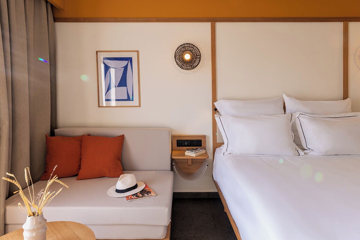 A hotel room with a bed, a sofa with orange pillows, a nightstand with a lamp, and a framed picture on the wall.