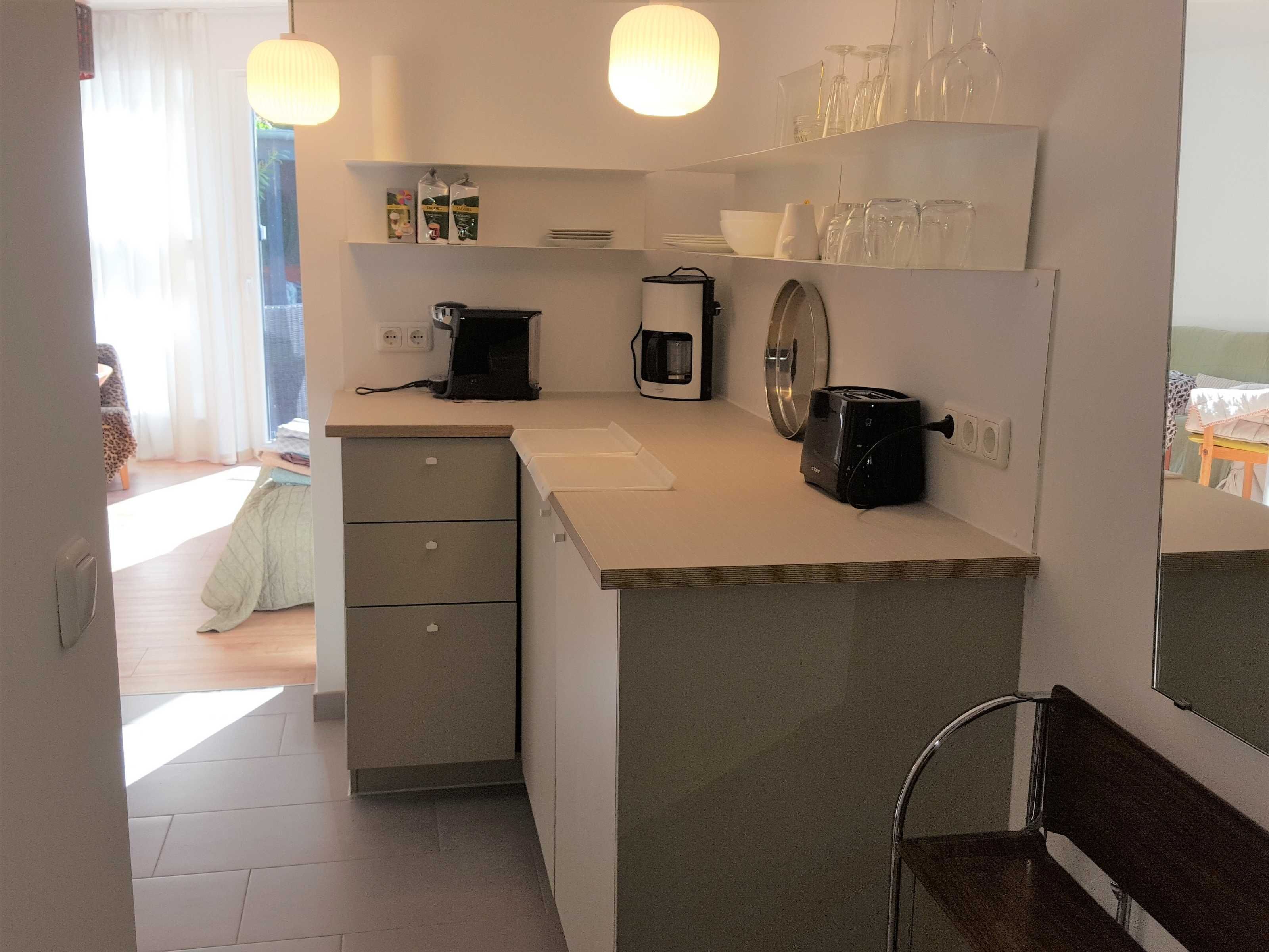 Eine kompakte Küche mit Kaffeemaschine, Toaster und kleinem Kühlschrank. Durch einen Durchgang ist ein Essbereich mit Tisch und Stühlen zu sehen.