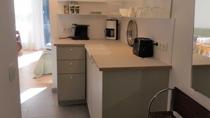 A compact kitchen with a coffee maker, toaster, and a small refrigerator. There is a dining area with a table and chairs visible through a doorway.