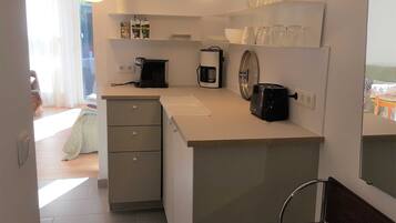 Eine kompakte Küche mit Kaffeemaschine, Toaster und kleinem Kühlschrank. Durch einen Durchgang ist ein Essbereich mit Tisch und Stühlen zu sehen.