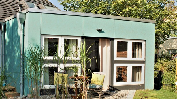 A modern house with a flat roof, large windows, and a small porch area with a chair and table.
