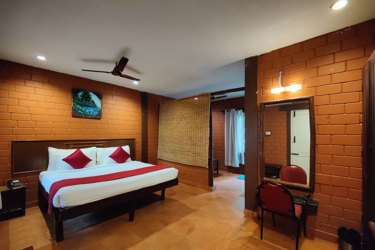 A hotel room with a brick wall, a bed with white linens and red pillows, a ceiling fan, a large mirror, and a wooden floor.