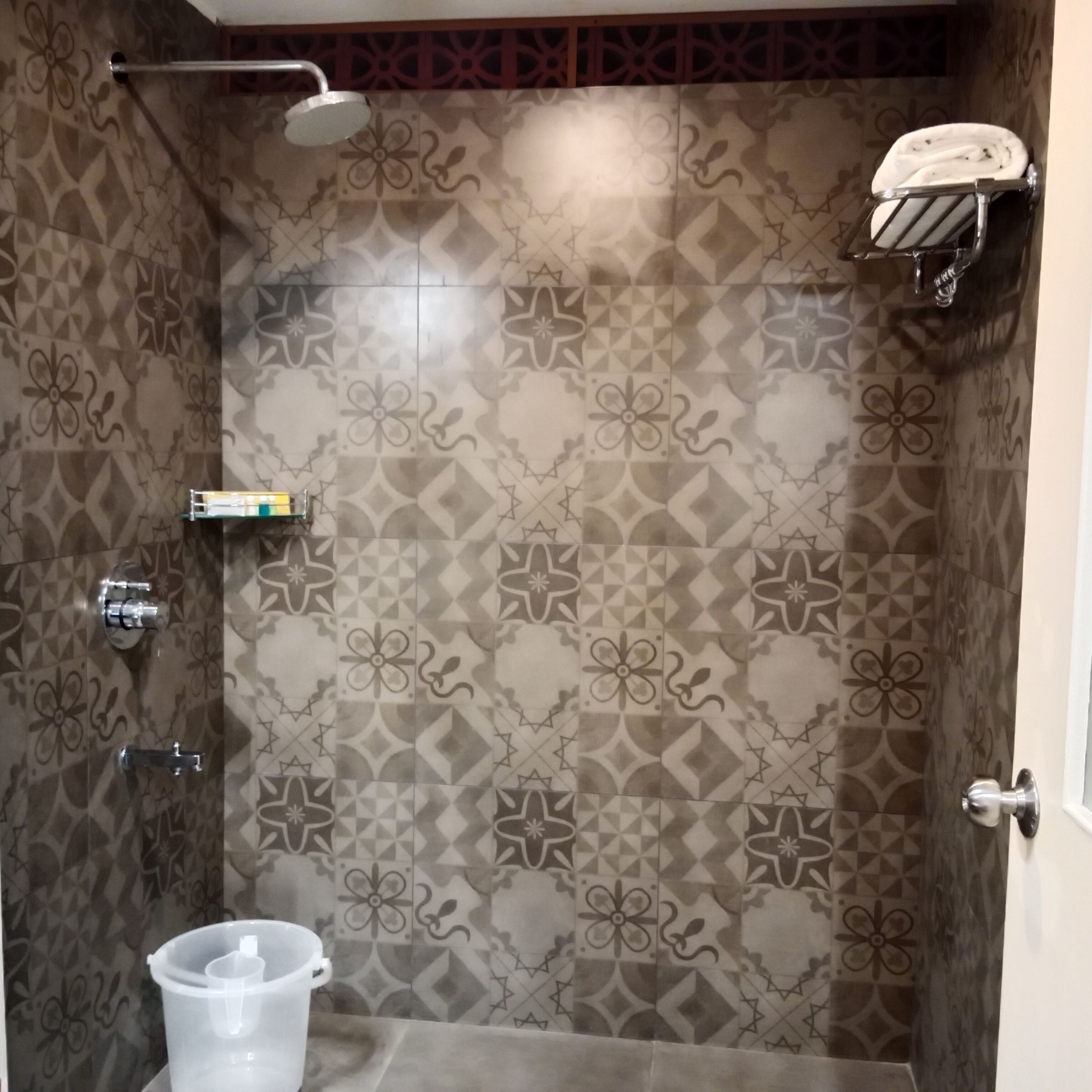 Zona de ducha con azulejos estampados, cabezal de ducha de mano, toallero y un cubo blanco.