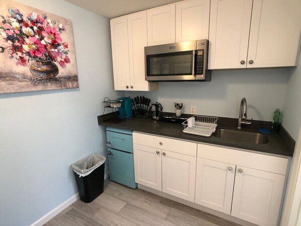 A kitchen with white cabinets, a stainless steel microwave, a sink, and a trash bin.
