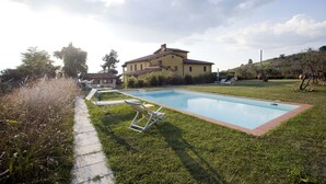 Una casa grande con piscina, rodeada de un césped cuidado y algunas sillas de descanso.