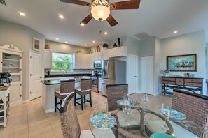 Una cocina moderna con armarios blancos, una isla central con asientos, un comedor con mobiliario de mimbre y un ventilador de techo.