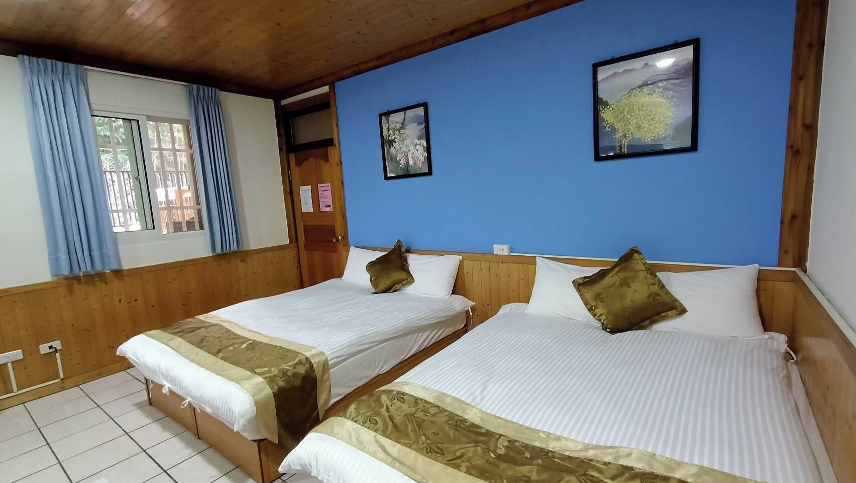 A room with two beds, wooden headboards, a blue wall, and a window with curtains.