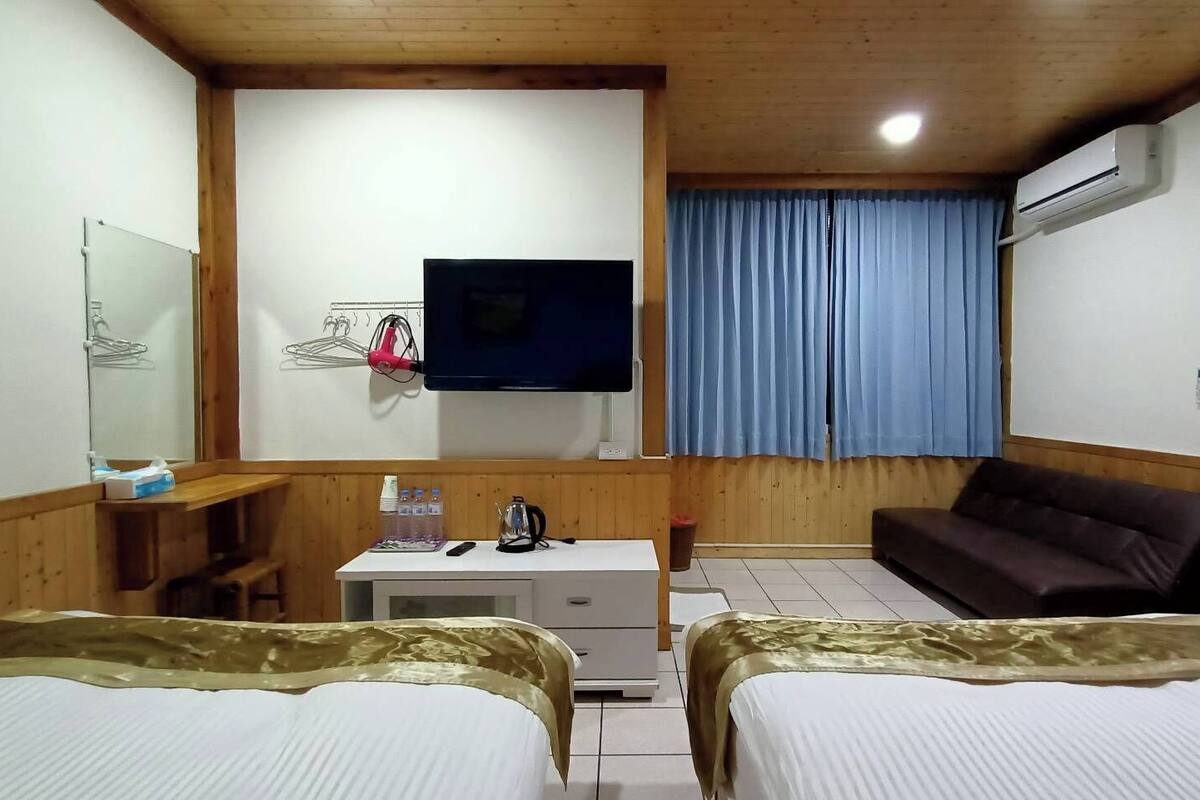 A hotel room with two beds, a desk with a flat-screen TV, a sofa, and a window with blue curtains.
