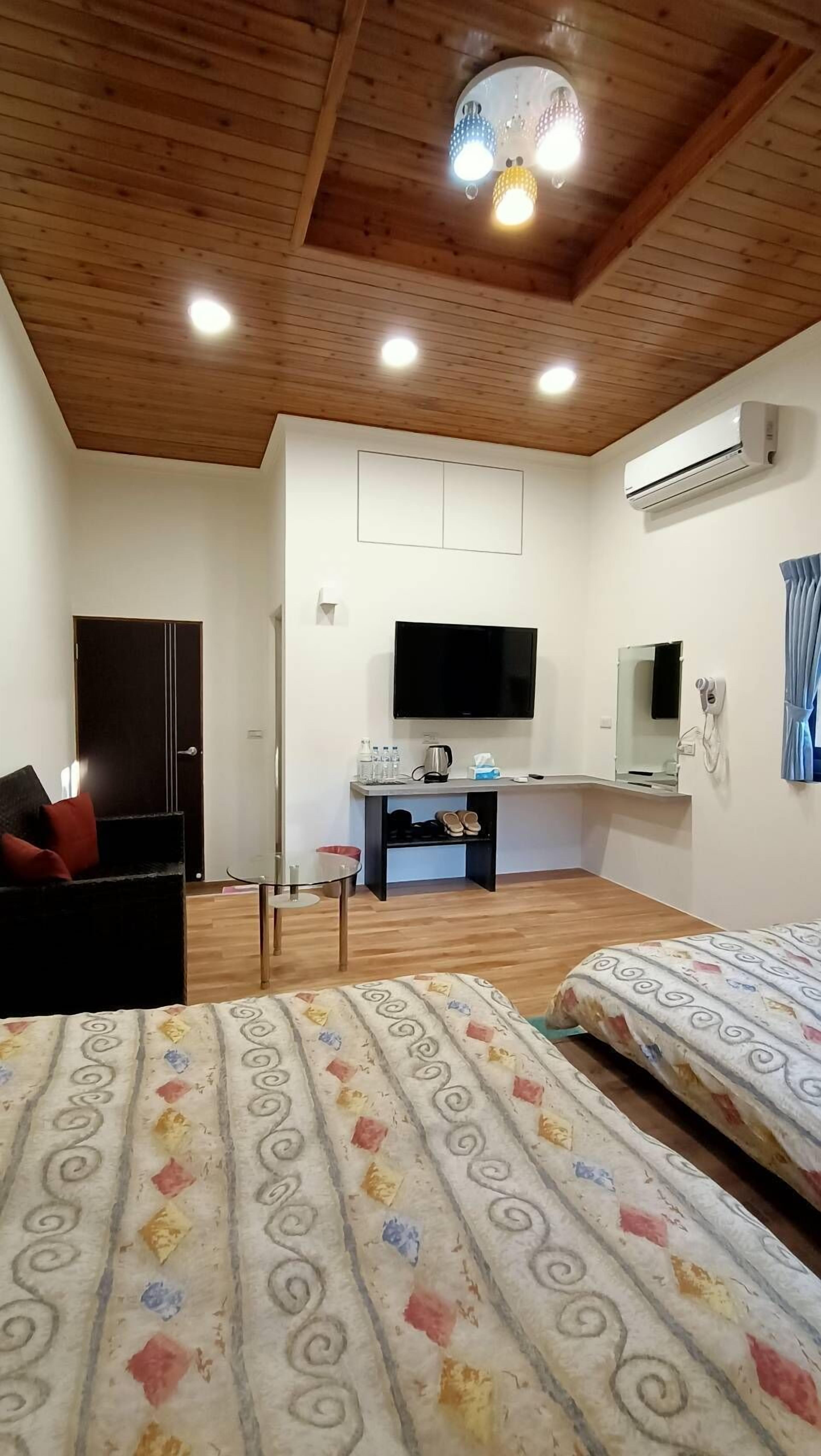 A hotel room with a wooden ceiling, a flat-screen TV, an air conditioner, a desk with a chair, a bed with patterned bedding, and a door.