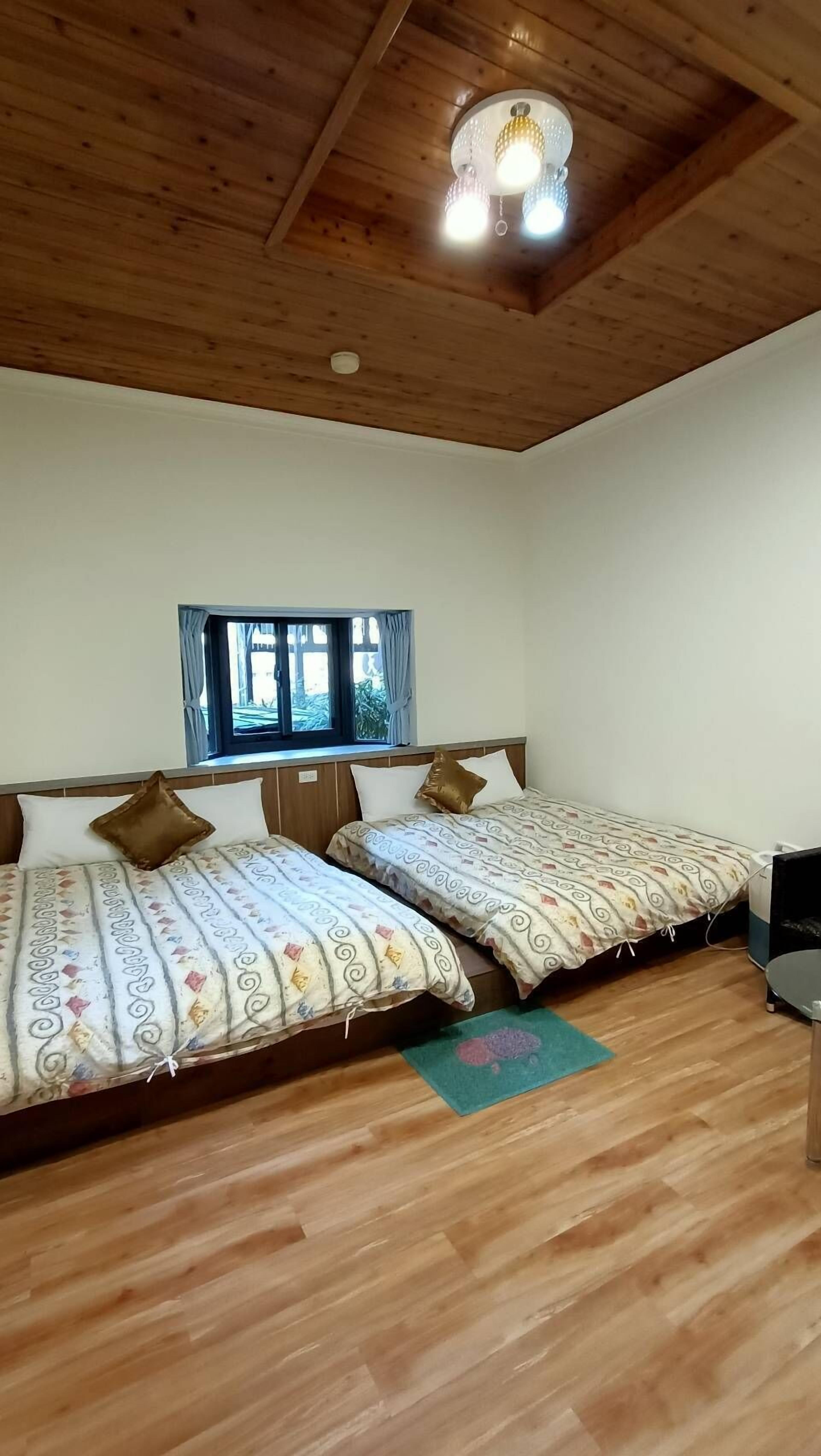 A room with two beds, wooden flooring, a window with curtains, and a ceiling light fixture.