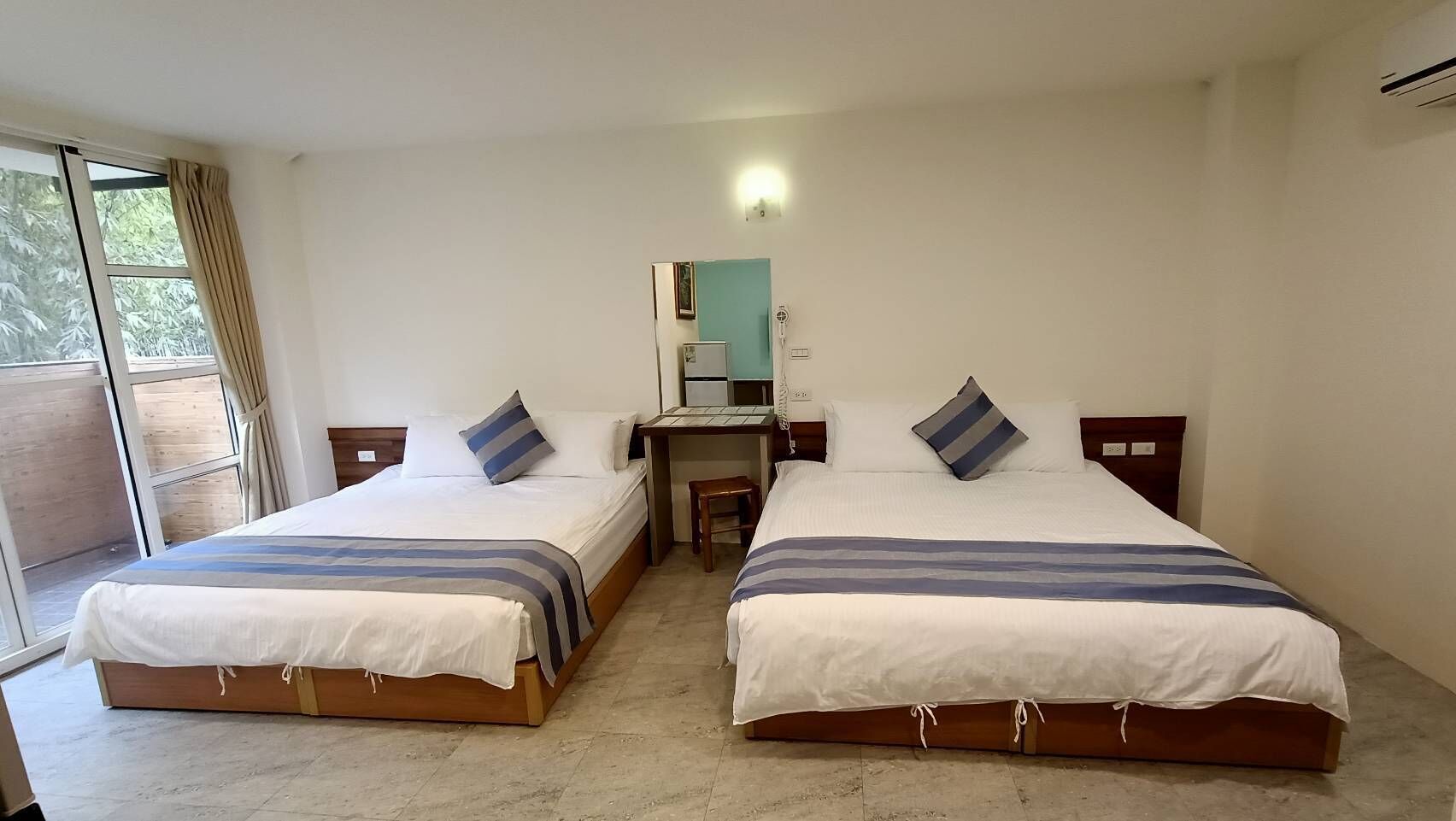 Une chambre d’hôtel avec deux lits simples, un petit bureau avec une chaise, un miroir et une fenêtre avec des rideaux.