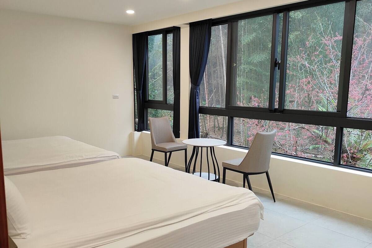 A hotel room with a large bed, two chairs, a small table, and a view of trees through large windows.