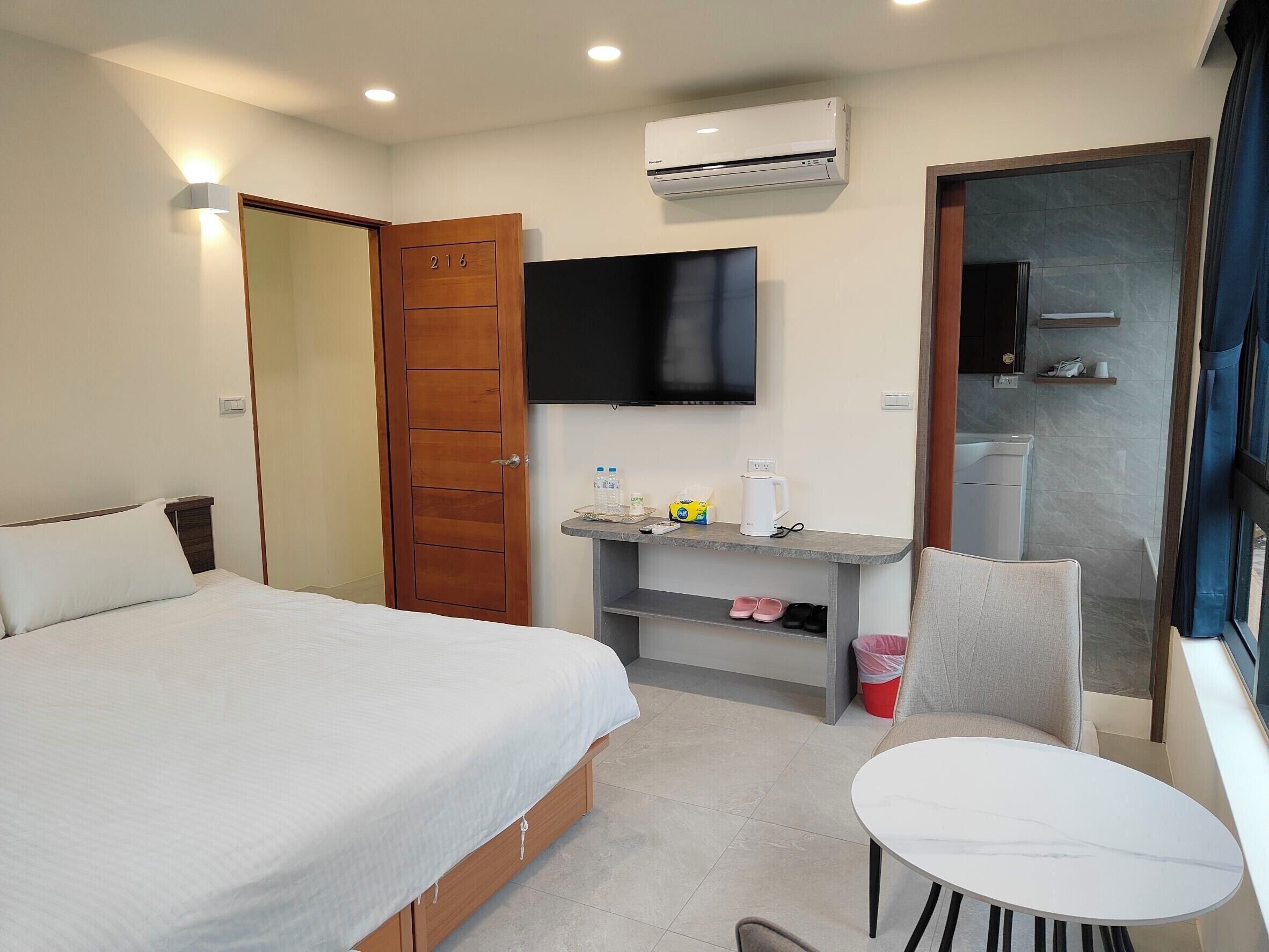 A hotel room with a bed, a desk with a flat-screen TV, a chair, and a small table.