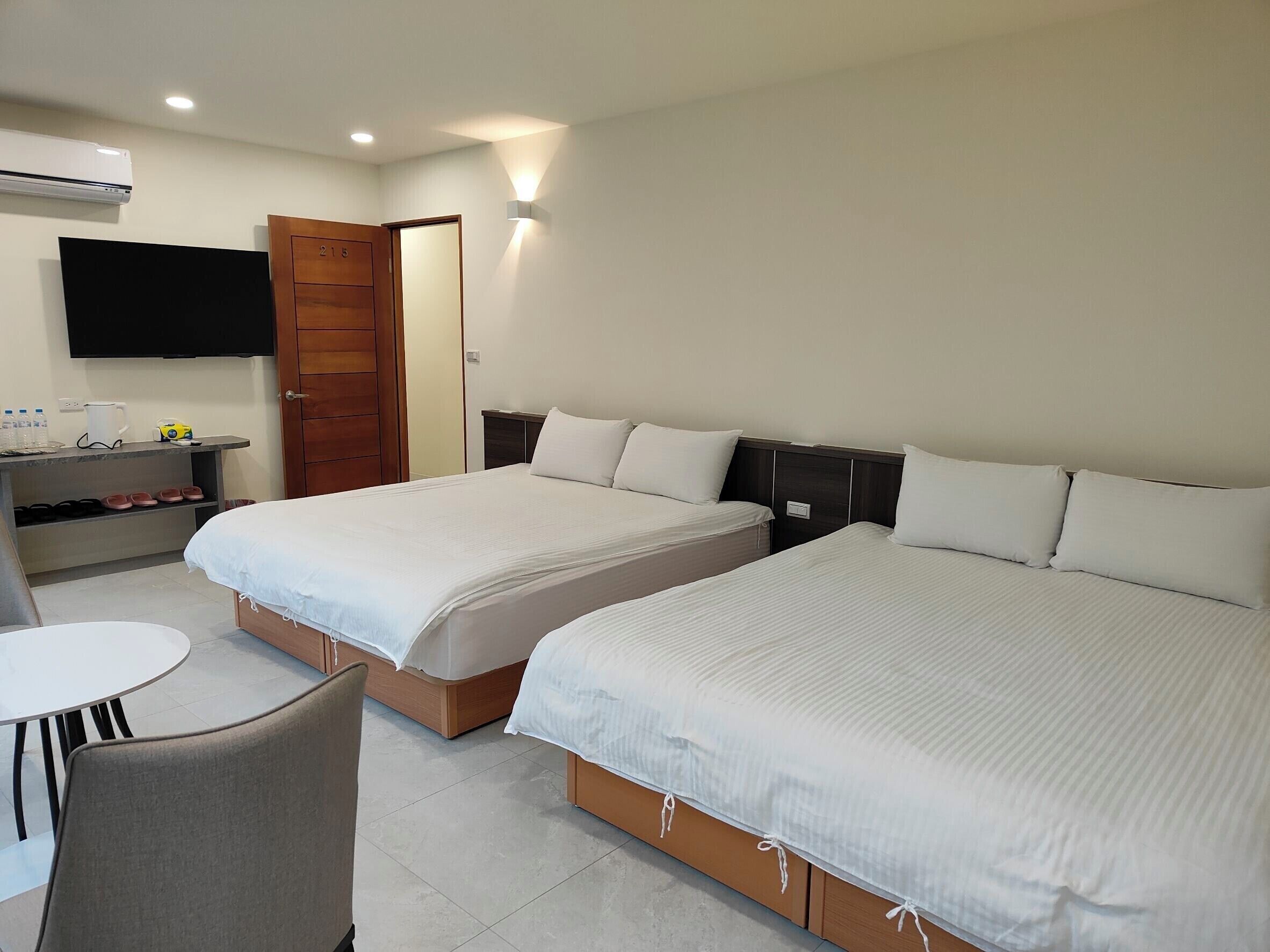 A hotel room with two beds, a desk with a chair, a flat-screen TV, and a door.