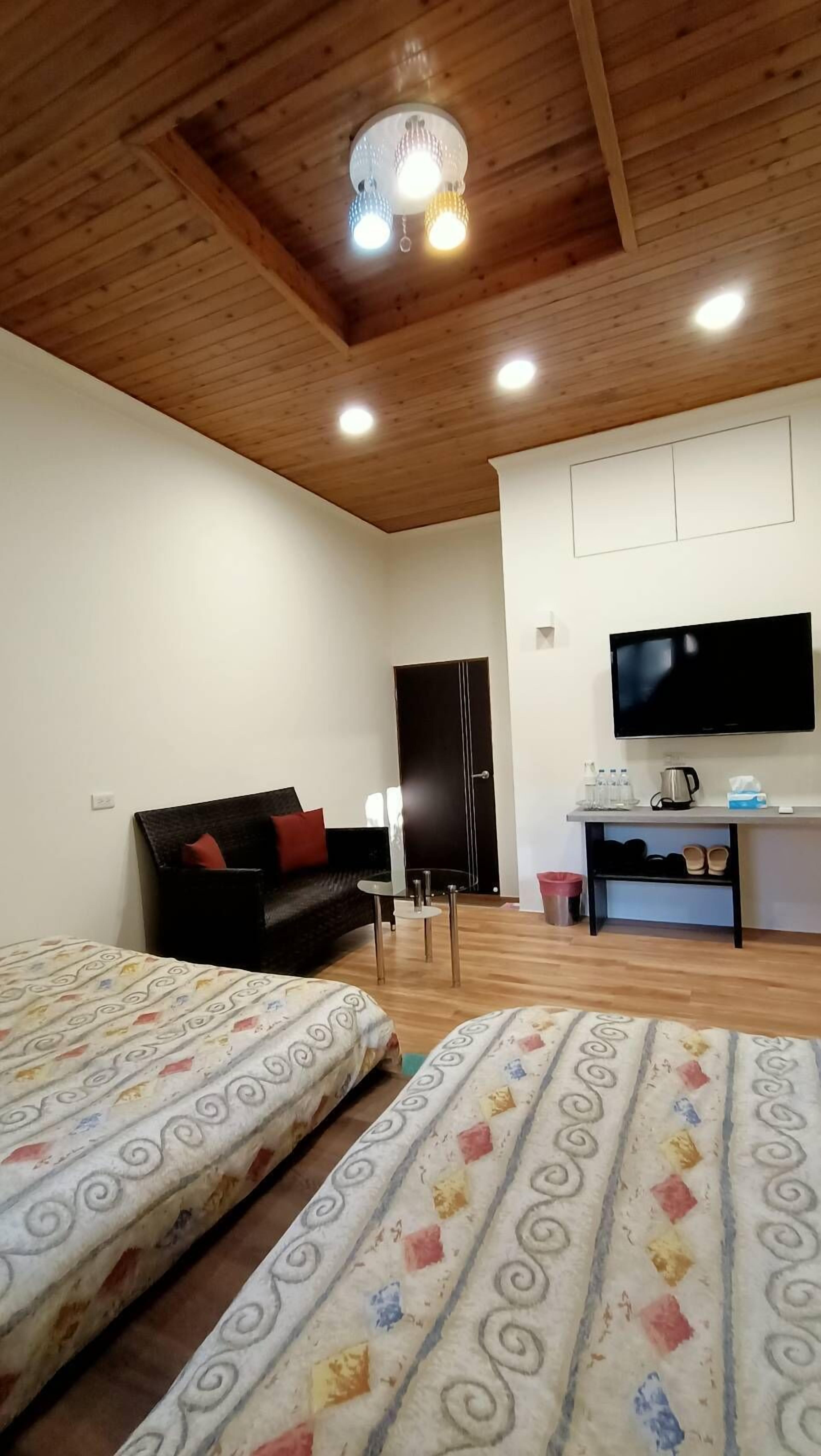 Une pièce avec des poutres apparentes en bois au plafond, un canapé noir avec des coussins rouges, deux lits avec une literie à motifs, un téléviseur fixé au mur et une petite table avec une bouilloire et des tasses.