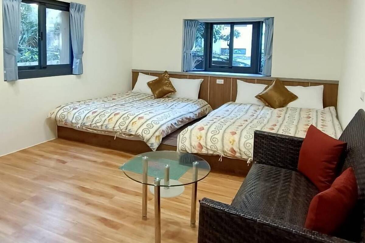 A room with two beds, a glass coffee table, a wicker sofa, wooden flooring, and a wooden ceiling with a light fixture.