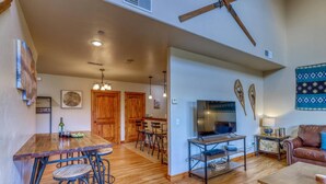 A spacious living area with a wooden dining table, bar stools, a TV, and a sofa. - Cozy Condo with Balcony, Walk to Shops, Dining, and Hot Springs Nearby! (Pagosa Springs)