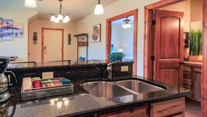 A kitchen with granite countertops, stainless steel sinks, and wooden cabinets. There are ceiling lights, a coffee maker, and a dish rack with dishes. - Cozy Condo with Balcony, Walk to Shops, Dining, and Hot Springs Nearby! (Pagosa Springs)