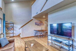 A spacious living area with a wooden floor, a leather sofa, a dining table with bar stools, a flat-screen TV, and a bookshelf.