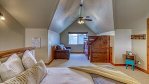 A loft bedroom with a sloped ceiling, a ceiling fan, a bed with a blanket, a sofa, a wooden staircase, and a window. - Cozy Condo with Balcony, Walk to Shops, Dining, and Hot Springs Nearby! (Pagosa Springs)