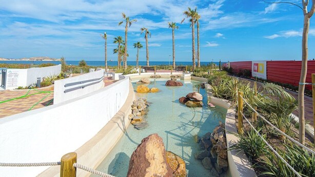A pool area with rocks and palm trees, a red building, and a view of the sea.