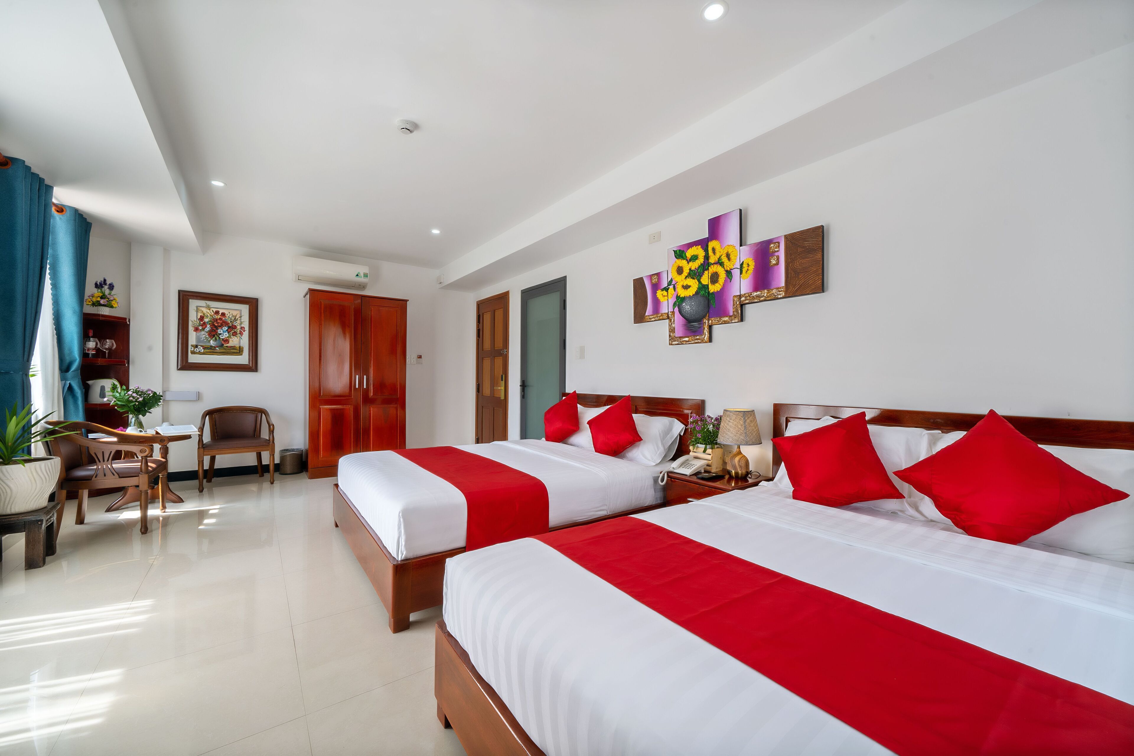 Ein Hotelzimmer mit zwei Betten, rot-weißer Bettwäsche, einem hölzernen Kopfteil, einem Schreibtisch mit Stuhl, einer Topfpflanze und einem an der Wand hängenden Kunstwerk.