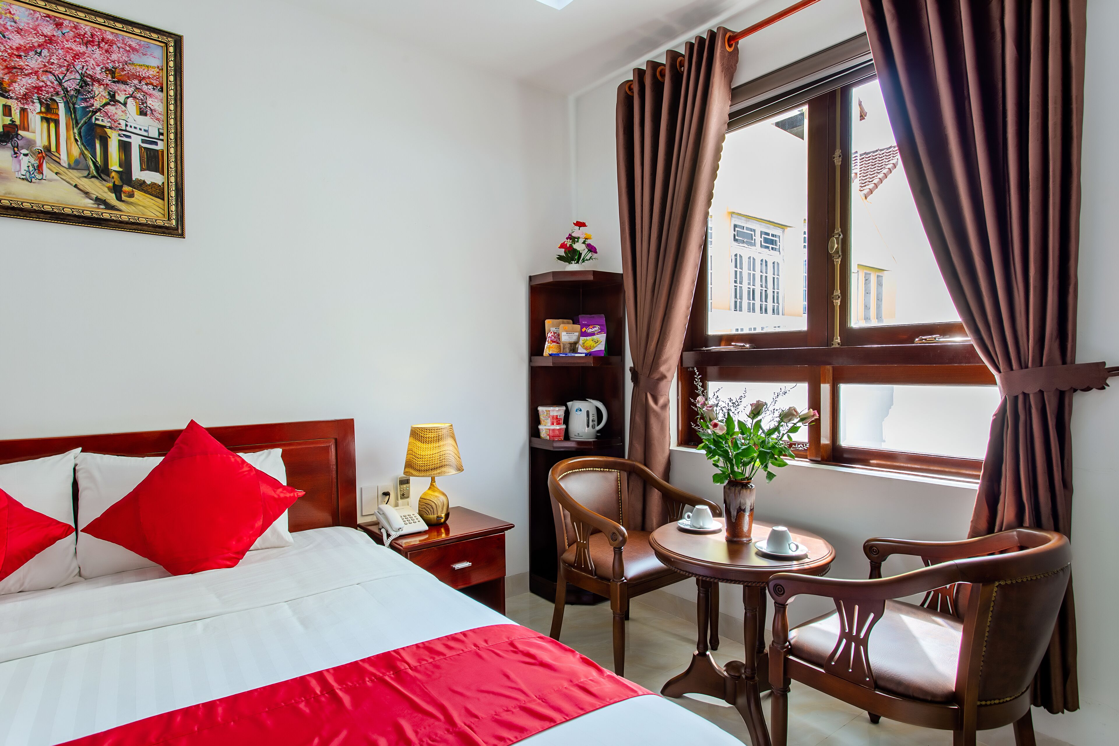 A hotel room with a bed, a desk, a chair, a small table with a vase, a bookshelf, and a window with curtains.