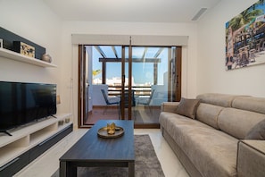 A modern living room with a flat-screen TV, a sofa, a coffee table, and a sliding glass door leading to a balcony with a view of the city. - Apartamento Neptuno (Mar de Pulpi) (San Juan de los terreros)