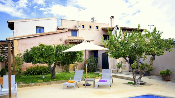 A pool area with lounge chairs and umbrellas, surrounded by a garden and a building with a tiled roof. - 44380 Finca Pula Viñas (Son Servera)