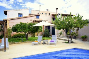 A pool area with lounge chairs and umbrellas, surrounded by a garden and a building with a tiled roof. - 44380 Finca Pula Viñas (Son Servera)