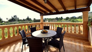A covered outdoor seating area with a round table and chairs, a balustrade, and a view of a green landscape. - 44380 Finca Pula Viñas (Son Servera)