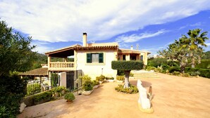 A Mediterranean-style villa with a tiled roof, a balcony, and a well-maintained garden with various plants and a palm tree. - 44380 Finca Pula Viñas (Son Servera)
