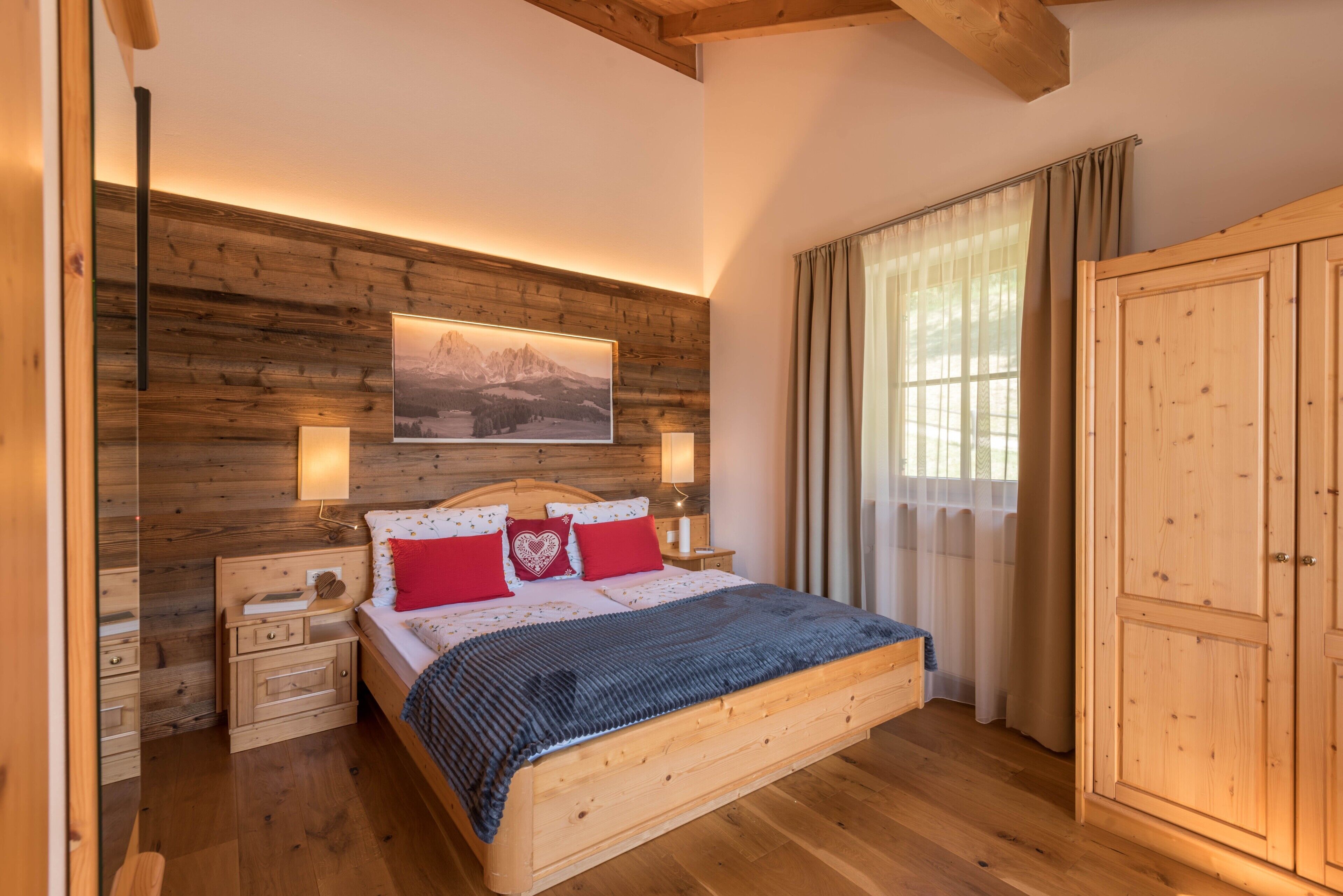 Ein Schlafzimmer mit Holzbettrahmen, einem Bett, Nachttischen, einem Kleiderschrank und einem großen Fenster mit Vorhängen.